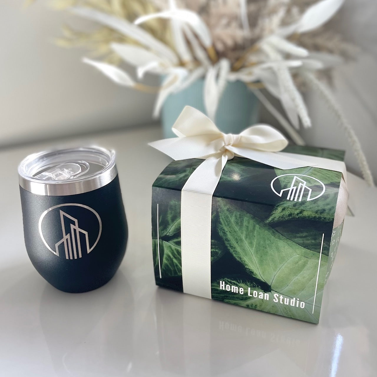 Black blue tumbler with silver lid next to a green box with white ribbon on a light surface.