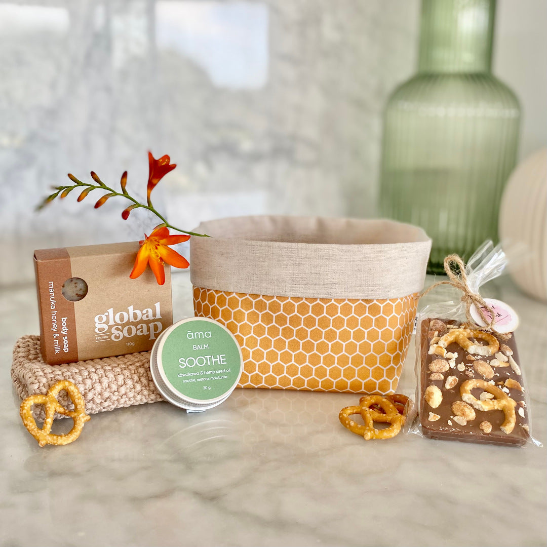 Gift Box NZ | New Zealand made products | Small gift boxes NZ | Gift in New Zealand | A gift box packed with Kiwi classics all packaged in a reusable honeycomb fabric box. Honey and milk body soap. Kawakawa balm Soothe in a tin. Pretzel & Peanut Milk Chocolate. Neutral knitted wash cloth. Honeycomb fabric gift box